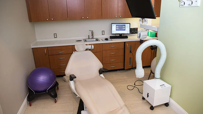 Modern dental treatment room with cream patient chair, wood cabinetry, computer monitor, intraoral camera, and air purification equipment